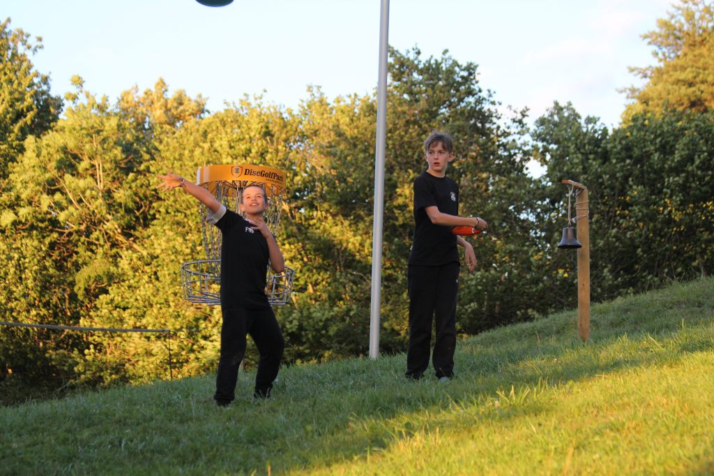Zwei Kinder spielen Discgolf. Im rechten Teil des Bildes ist die Essensglocke zu sehen