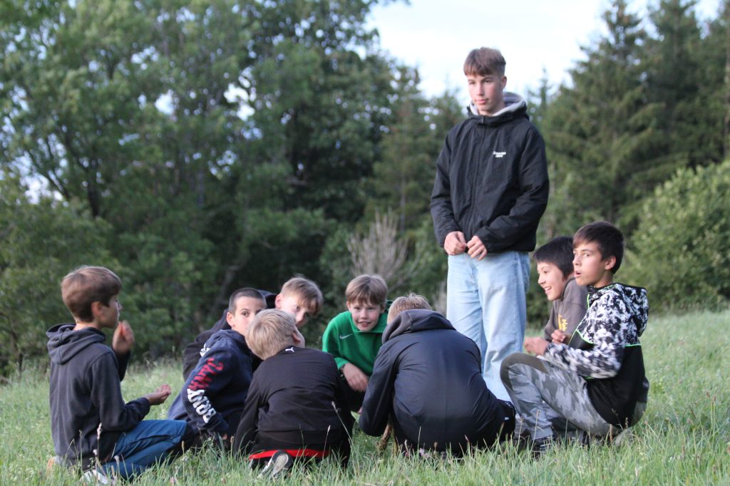 Eine Gruppe von Kindern sitzt in einem Kreis auf einer Wiese. Ein Mitarbeiter steht in ihrer Mitte