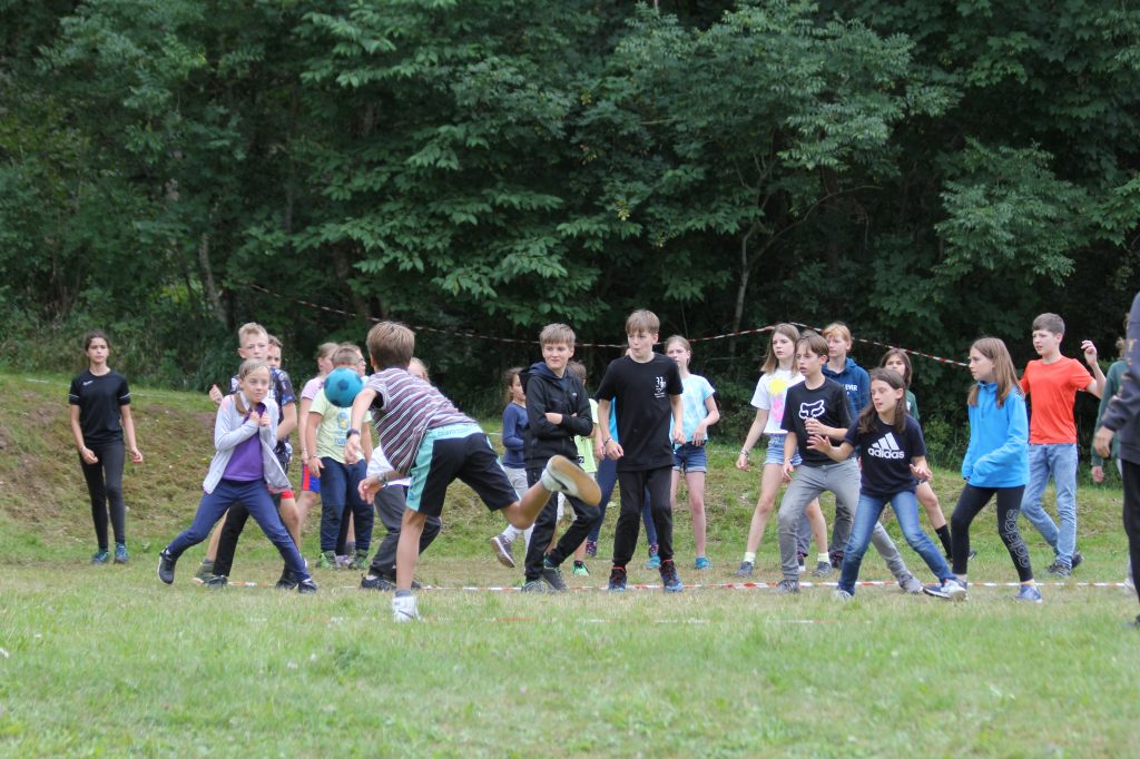 Sehr viele Kinder stehen in einem mit Flatterband abgegrenzten Völkerballfeld. Ein Schaumstoffball fliegt in Richtung der Menge